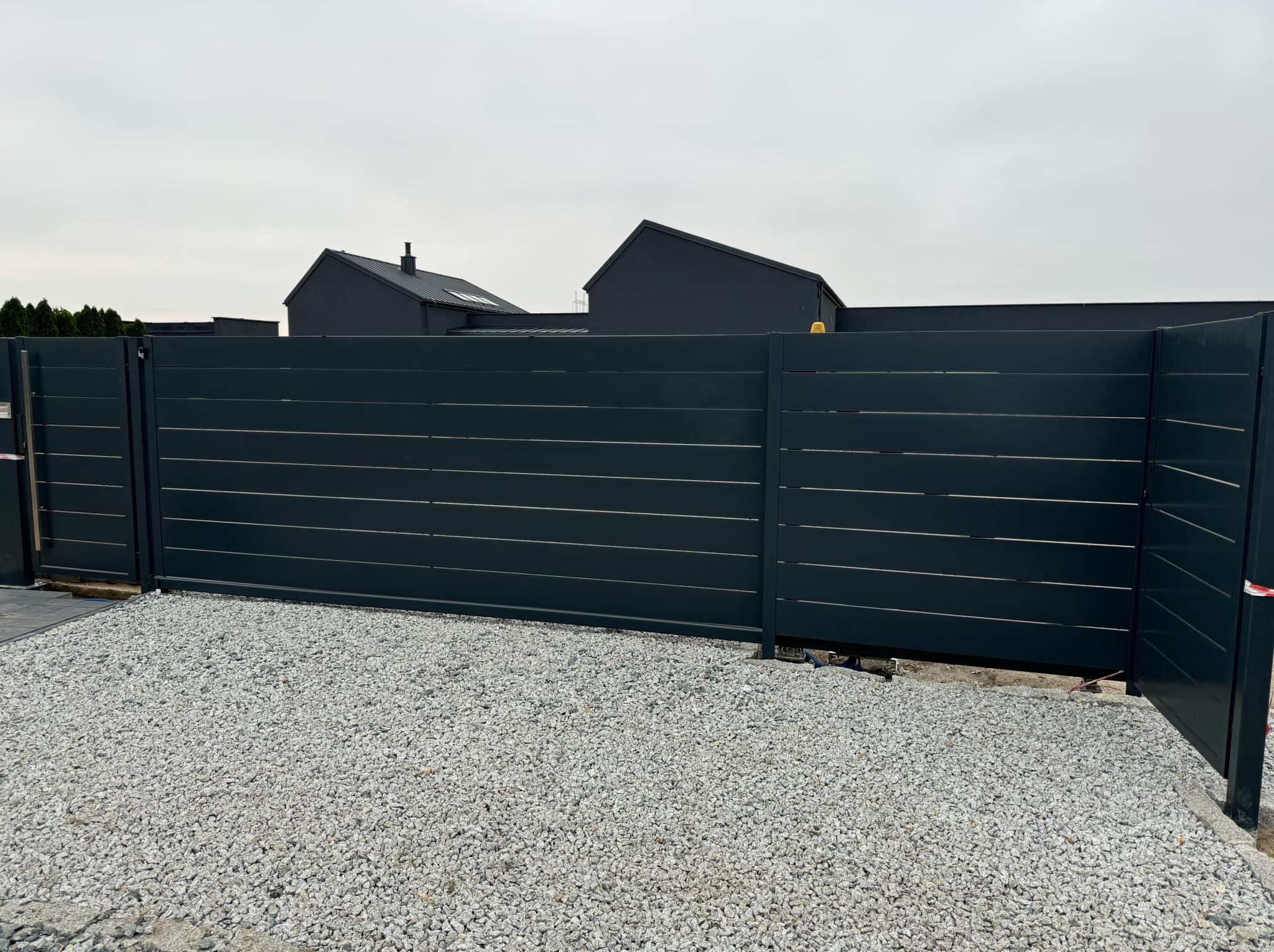 Nowoczesna, przesuwna brama wjazdowa z poziomymi panelami w kolorze grafitowym na tle szarego nieba i domu o minimalistycznej architekturze, wysypana żwirem.