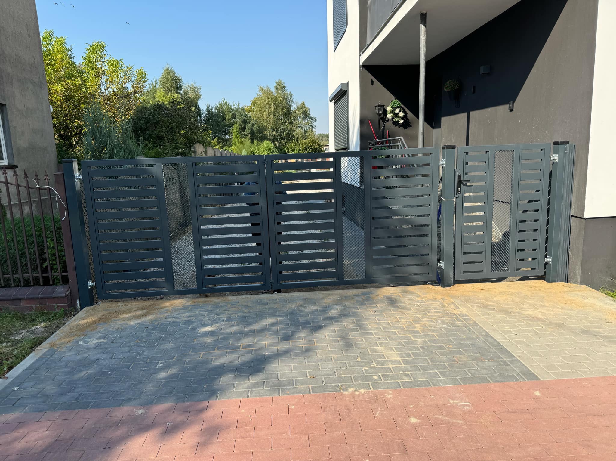 Nowoczesna, grafitowa brama wjazdowa z poziomymi szczeblami, uzupełniona kratką. Brama zamontowana na posesji z kostką brukową, z domem w tle.