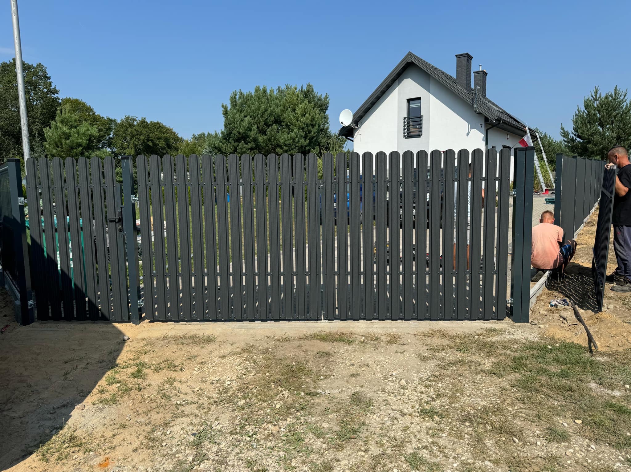 Grafitowa brama wjazdowa z pionowych sztachet, częściowo zamontowana. W tle dom z białą elewacją i ciemnym dachem, fragment sylwetki pracownika.