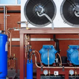 Kompresory klimatyzacji przemysłowej na zewnątrz budynku z cegły, widoczne rury i instalacje. Dwa duże wentylatory na górze.