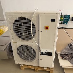 AGDKlinika - Biała jednostka zewnętrzna klimatyzacji Copeland Scroll na palecie w surowym wnętrzu. Widoczne narzędzia i materiały budowlane w tle. Emerson.