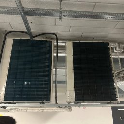 AGDKlinika - Dwa duże, zamontowane pod sufitem klimatyzatory przemysłowe z widocznymi przewodami i instalacją. Metalowe korytka kablowe pod sufitem.