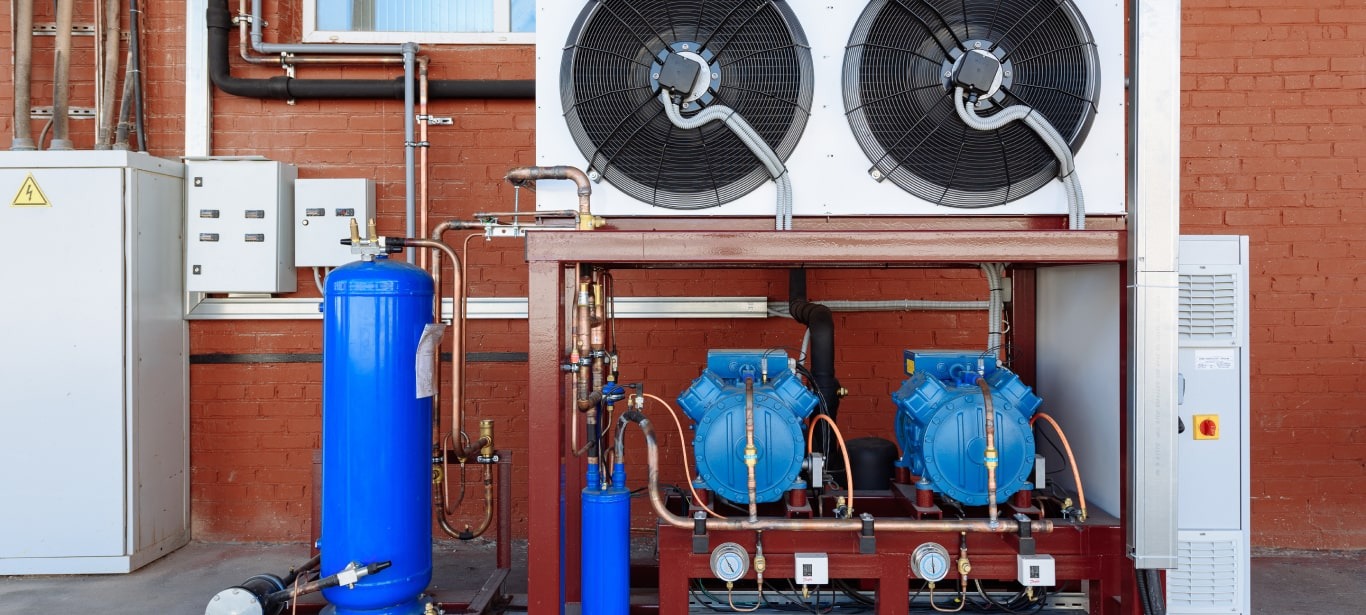 Kompresory klimatyzacji przemysłowej na zewnątrz budynku z cegły, widoczne rury i instalacje. Dwa duże wentylatory na górze.