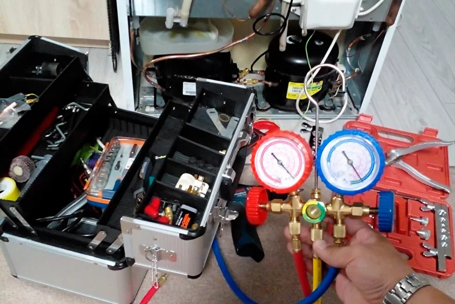 Otwarta walizka narzędziowa obok lodówki w trakcie naprawy. Mierniki ciśnienia w dłoni serwisanta. Widoczne elementy instalacji chłodniczej.