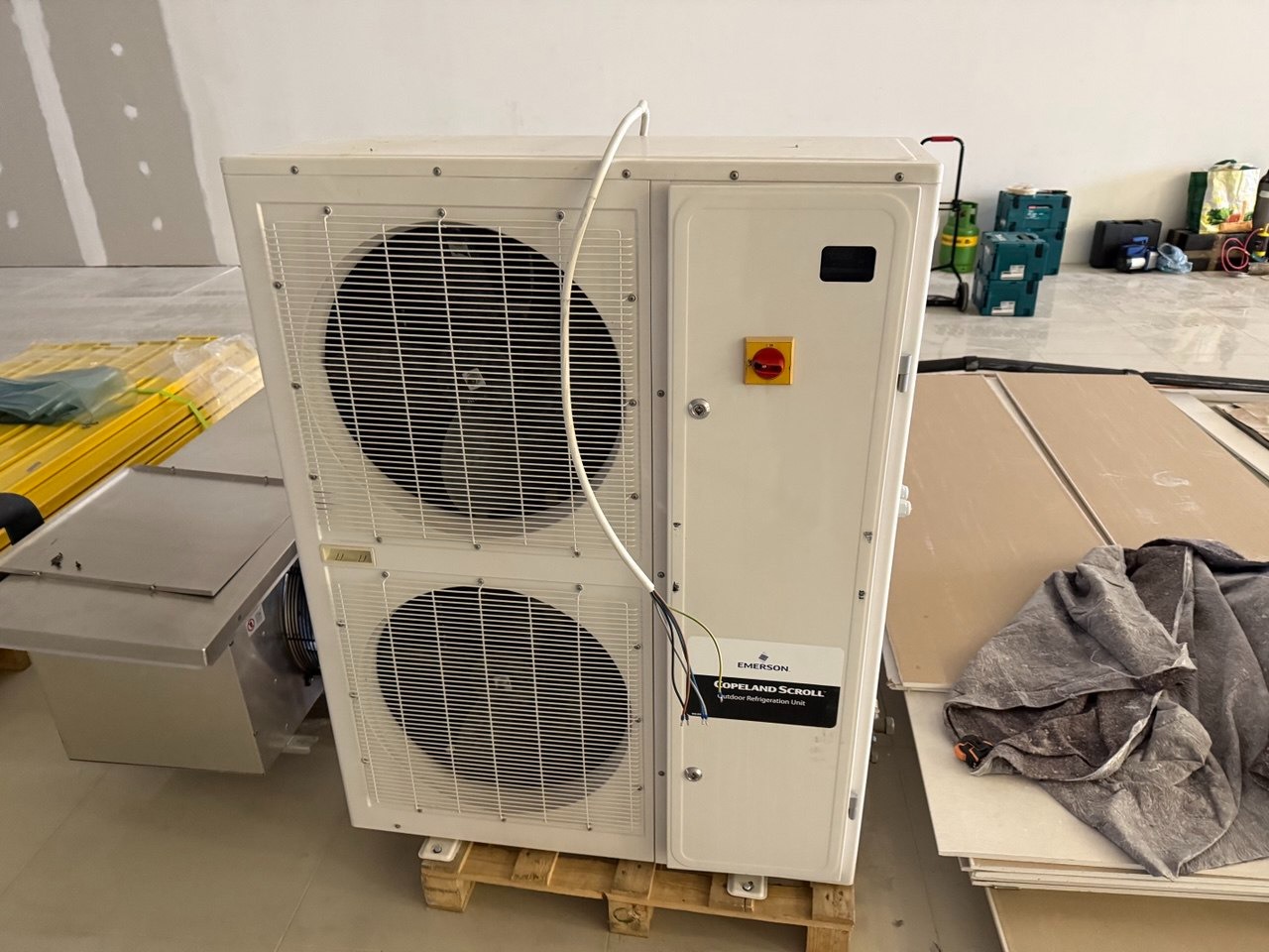 Biała jednostka zewnętrzna klimatyzacji Copeland Scroll na palecie w surowym wnętrzu. Widoczne narzędzia i materiały budowlane w tle. Emerson.