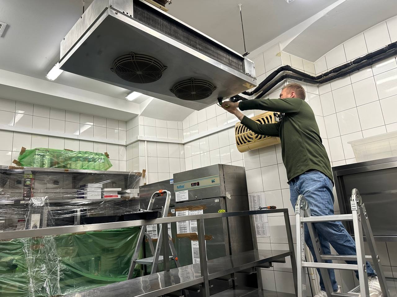 Mężczyzna na drabinie serwisuje przemysłową klimatyzację sufitową w lokalu gastronomicznym. Widoczne metalowe elementy, wentylatory i instalacja.