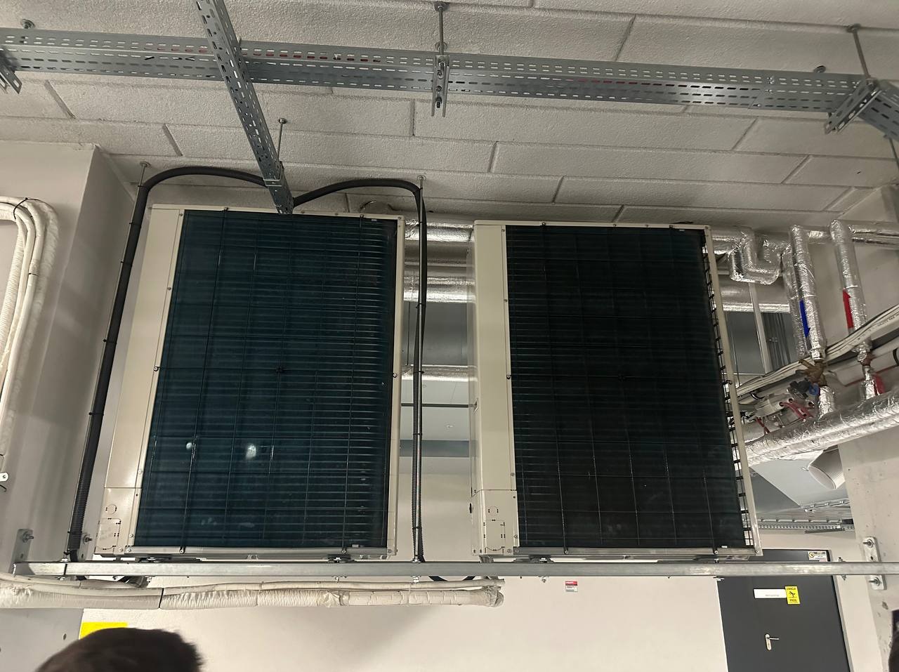 Dwa duże, zamontowane pod sufitem klimatyzatory przemysłowe z widocznymi przewodami i instalacją. Metalowe korytka kablowe pod sufitem.