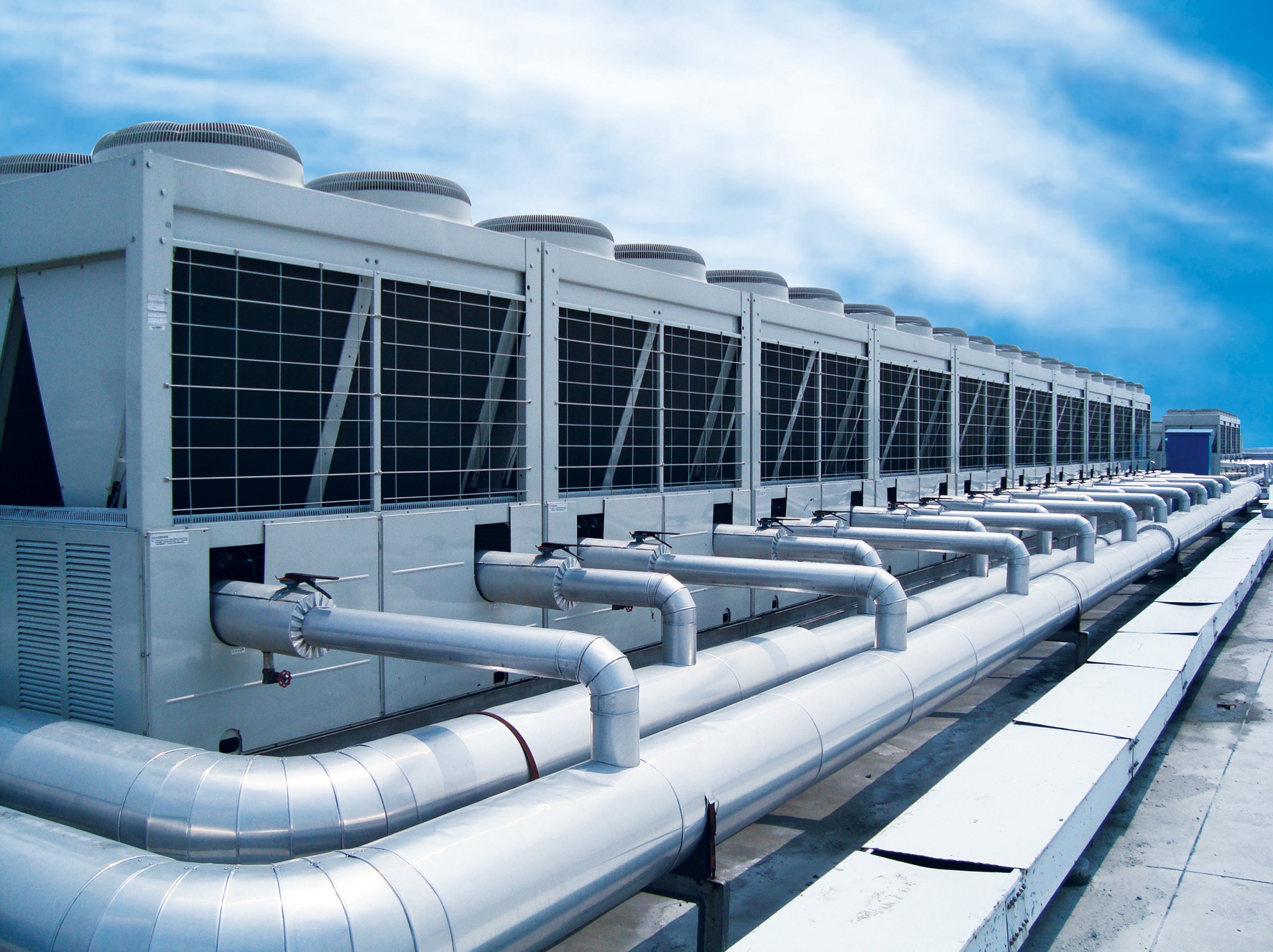 Szereg przemysłowych klimatyzatorów na dachu budynku z widocznymi rurami, na tle błękitnego nieba z chmurami. Widok z dołu, podkreślający skalę instalacji.