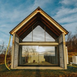 AS BUD DEVELOPMENT SPÓŁKA Z OGRANICZONĄ ODPOWIEDZIALNOŚCIĄ - Surowy stan domu z dużym przeszkleniem i drewnianą konstrukcją dachu, otoczony zielenią i elementami budowy, uchwycony w pogodny dzień.