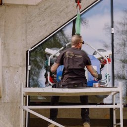 AS BUD DEVELOPMENT SPÓŁKA Z OGRANICZONĄ ODPOWIEDZIALNOŚCIĄ - Montaż okna o nietypowym kształcie w surowym domu. Pracownik na rusztowaniu używa przyssawki i dźwigu. Widoczne bloczki z betonu komórkowego.