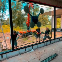AS BUD DEVELOPMENT SPÓŁKA Z OGRANICZONĄ ODPOWIEDZIALNOŚCIĄ - Montaż okna panoramicznego z użyciem dźwigu przyssawkowego. Trzech monterów precyzyjnie ustawia szybę w surowym wnętrzu domu w trakcie budowy.