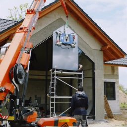 AS BUD DEVELOPMENT SPÓŁKA Z OGRANICZONĄ ODPOWIEDZIALNOŚCIĄ - Montaż okna na elewacji domu jednorodzinnego przy użyciu dźwigu z przyssawkami. Dwóch pracowników na rusztowaniu. Widoczny fragment ocieplonej ściany i dach.