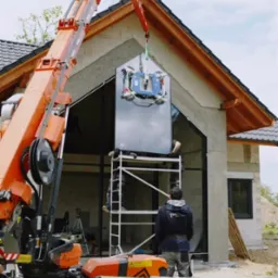 Montaż okna na elewacji domu jednorodzinnego przy użyciu dźwigu z przyssawkami. Dwóch pracowników na rusztowaniu. Widoczny fragment ocieplonej ściany i dach.