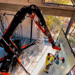 AS BUD DEVELOPMENT SPÓŁKA Z OGRANICZONĄ ODPOWIEDZIALNOŚCIĄ - Montaż okna w surowym budynku z użyciem robota-dźwigu BG Lift. Pracownik w odblaskowej kamizelce przykłada szybę. Widok z góry na plac budowy.