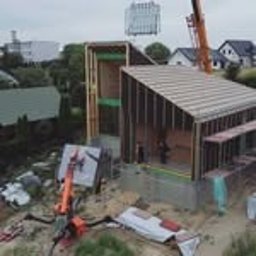 AS BUD DEVELOPMENT SPÓŁKA Z OGRANICZONĄ ODPOWIEDZIALNOŚCIĄ - Montaż domu szkieletowego: dźwig unosi gotowy moduł ściany z oknami. Widoczna konstrukcja domu w trakcie budowy, z zieloną izolacją. Dom w otoczeniu zieleni.