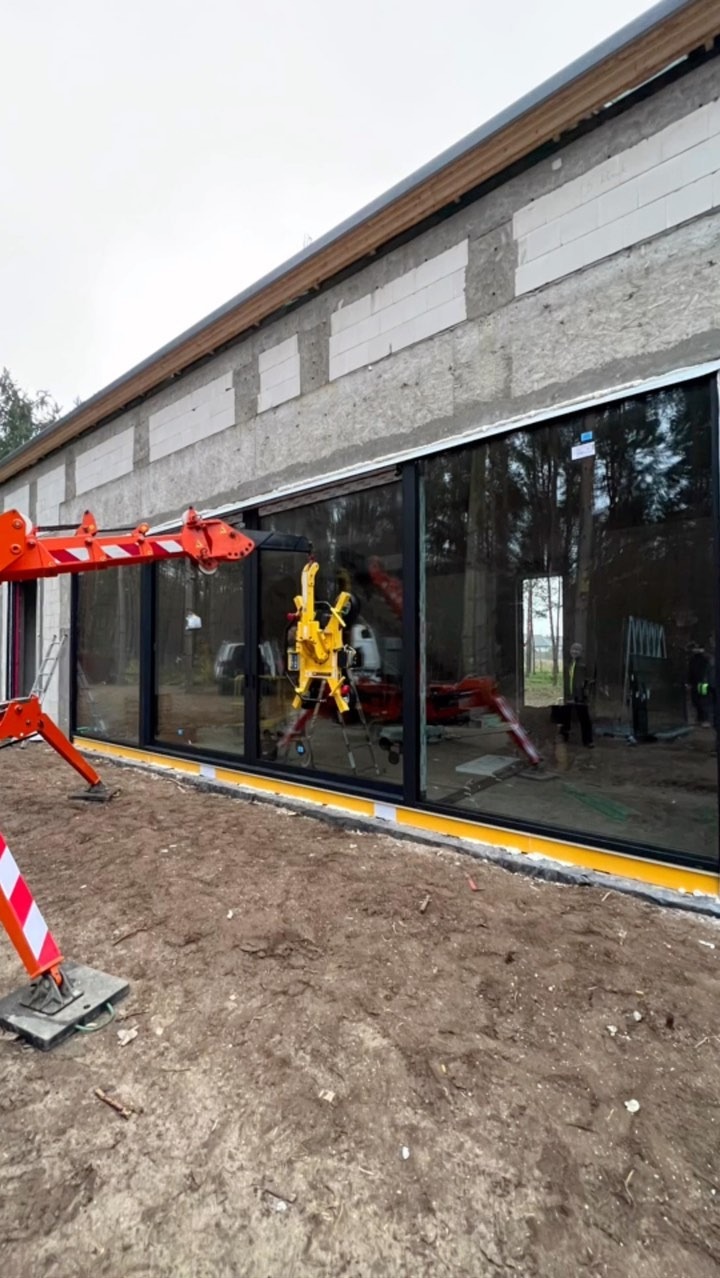 Montaż dużych okien przesuwnych w surowym budynku z betonu komórkowego, z użyciem specjalistycznego dźwigu. Widoczne elementy konstrukcyjne i żółta taśma zabezpieczająca.