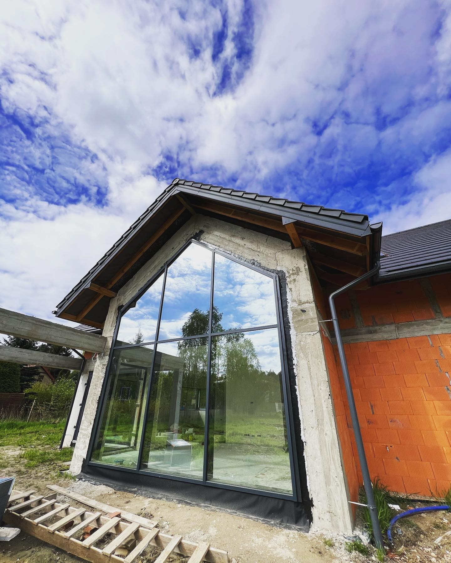 Surowy stan domu z dużym, trójkątnym oknem, w którym odbija się błękitne niebo. Widoczne cegły i beton, dach pokryty dachówką, drewniane elementy konstrukcyjne.