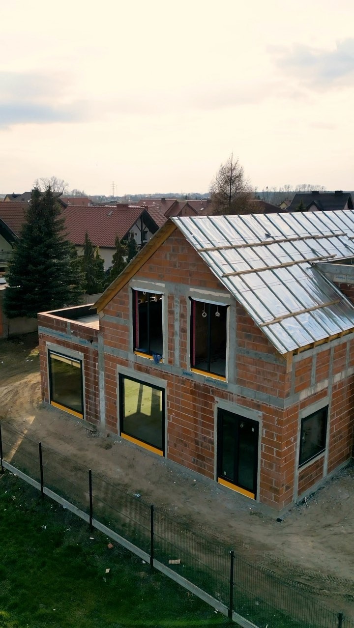 Ceglany dom w stanie surowym otwartym, z czarnymi oknami i zabezpieczonym dachem folią, na tle zabudowy jednorodzinnej i pochmurnego nieba.