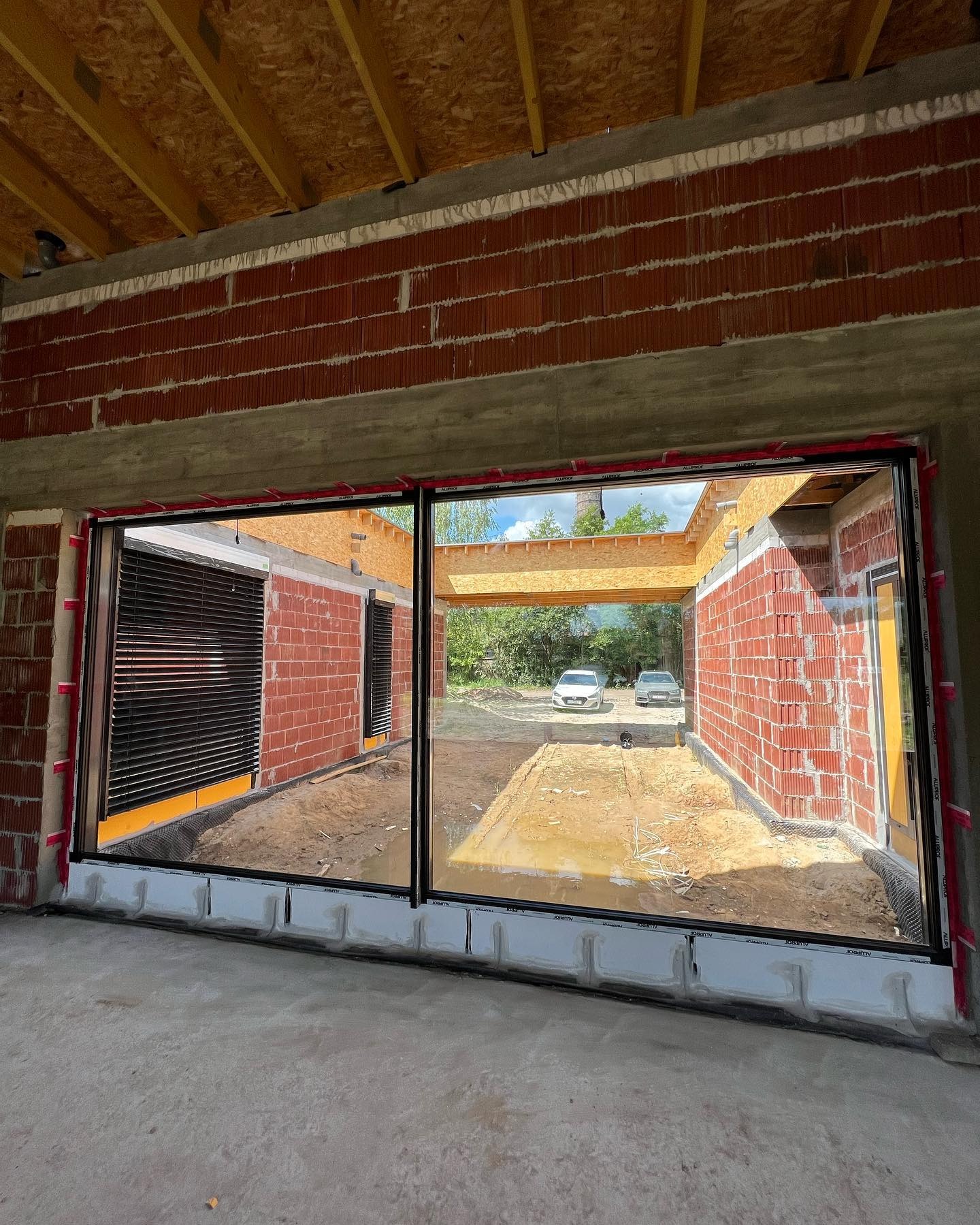 Wnętrze surowego domu z czerwonej cegły z dużym oknem. Widok na podjazd i dwa samochody. Konstrukcja dachu z desek. Elementy izolacji okiennej.
