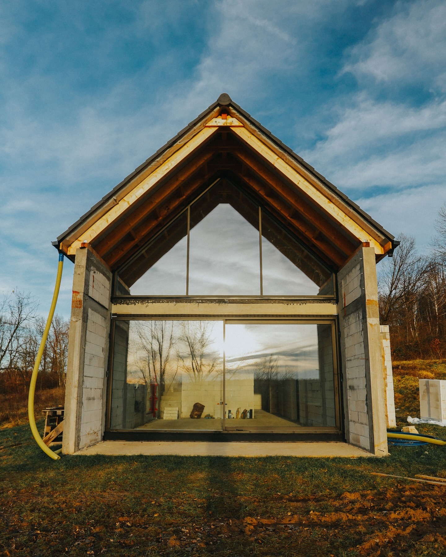 Surowy stan domu z dużym przeszkleniem i drewnianą konstrukcją dachu, otoczony zielenią i elementami budowy, uchwycony w pogodny dzień.