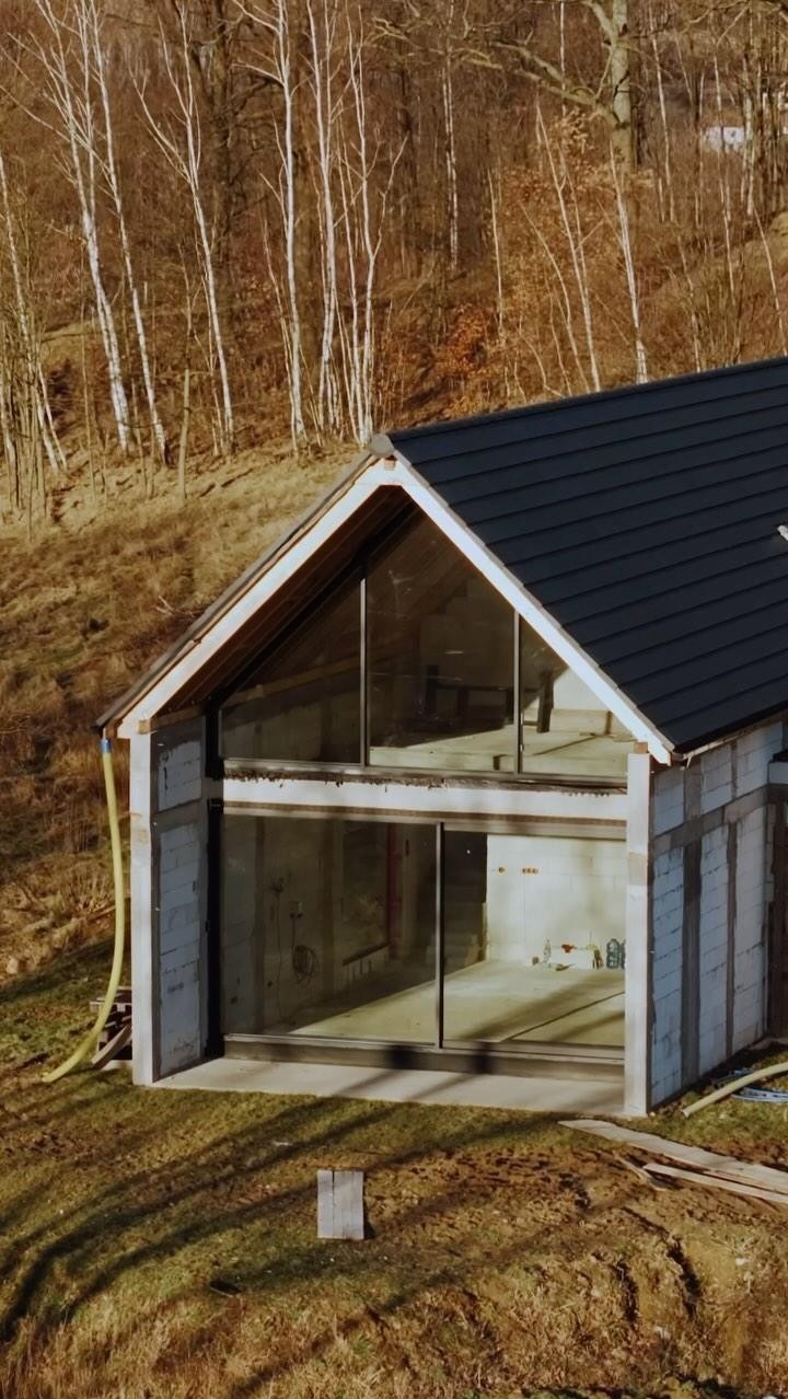 Niedokończony dom z bloków, z dużymi oknami i czarnym dachem na tle wzgórza z drzewami. Widoczna żółta rura i drewniane deski. Etap budowy, stan surowy otwarty.