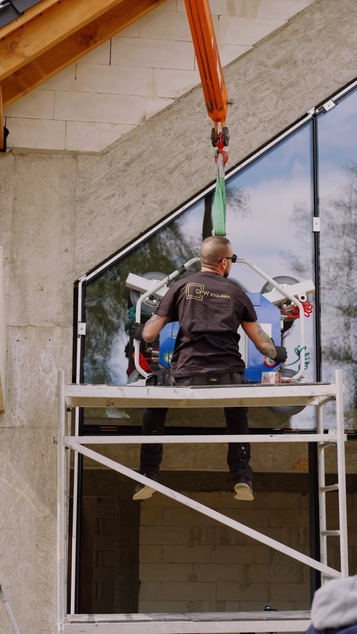 Montaż okna o nietypowym kształcie w surowym domu. Pracownik na rusztowaniu używa przyssawki i dźwigu. Widoczne bloczki z betonu komórkowego.