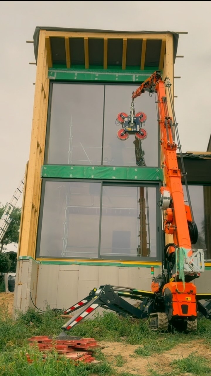 Montaż okien w domu szkieletowym za pomocą specjalistycznego dźwigu z przyssawkami. Widoczne elementy konstrukcji i izolacji.