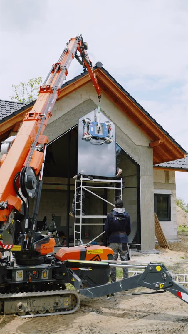 Montaż okna na elewacji domu jednorodzinnego przy użyciu dźwigu z przyssawkami. Dwóch pracowników na rusztowaniu. Widoczny fragment ocieplonej ściany i dach.