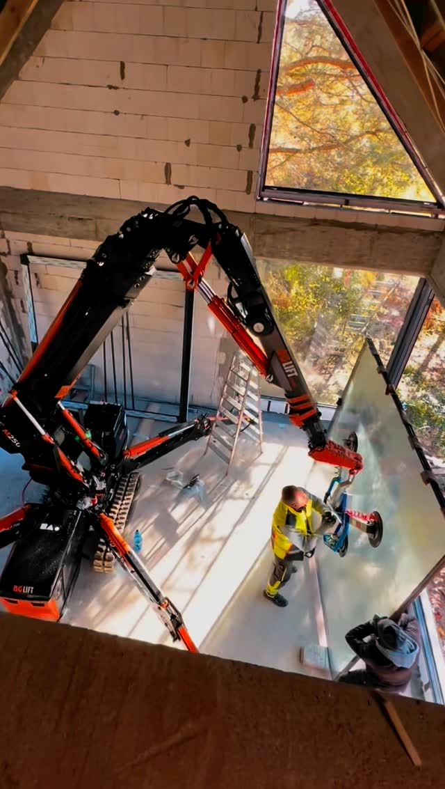 Montaż okna w surowym budynku z użyciem robota-dźwigu BG Lift. Pracownik w odblaskowej kamizelce przykłada szybę. Widok z góry na plac budowy.