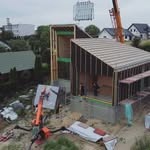 Montaż domu szkieletowego: dźwig unosi gotowy moduł ściany z oknami. Widoczna konstrukcja domu w trakcie budowy, z zieloną izolacją. Dom w otoczeniu zieleni.