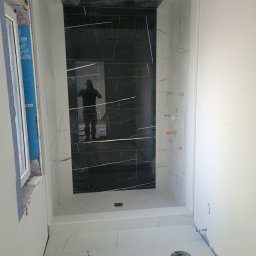 Soldatke Łazienki - Nowoczesna kabina prysznicowa z czarnymi płytkami imitującymi marmur i białymi płytkami podłogowymi. Widoczne elementy instalacji i odbicie osoby w tafli płytek.