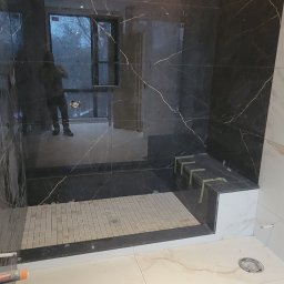 Soldatke Łazienki - Wykończona kabina prysznicowa z czarnymi płytkami imitującymi marmur i jasną mozaiką na brodziku. Widoczne jasne płytki podłogowe i odbicie osoby w szkle.