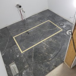 Soldatke Łazienki - Ułożona podłoga z ciemnych płytek imitujących marmur w łazience. Widoczne taśmy wyznaczające obszar, przygotowane podejście wod-kan i odpływ liniowy.