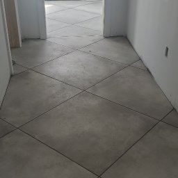 Soldatke Łazienki - Ukończona podłoga z szarych, kwadratowych płytek ułożonych pod kątem. Widoczne przewody elektryczne. W tle białe drzwi i ściany w trakcie wykańczania.