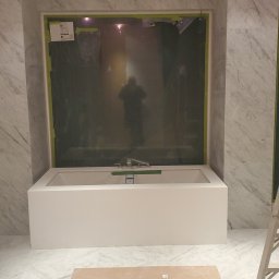 Soldatke Łazienki - Biała wanna obudowana marmurową ścianą w trakcie remontu łazienki. Zabezpieczone lustro, podłoga wyłożona marmurem, widoczna drabina i płyta zabezpieczająca podłogę.