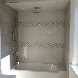 Soldatke Łazienki - Wykończona łazienka: biała wanna pod prysznicem z szarą mozaiką w zygzaki na jasnych płytkach. Widoczny panel boczny wanny i chromowana armatura.