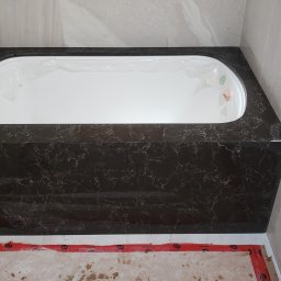 Soldatke Łazienki - Obudowana wanna z ciemnego marmuru z białym wnętrzem, zabezpieczona folią. W tle jasne, marmurowe płytki i fragment podłogi w trakcie remontu.