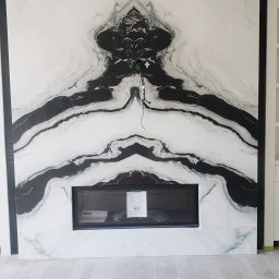 Soldatke Łazienki - Ściana z marmurowymi płytami z czarnym wzorem nad kominkiem. Widoczne kable elektryczne. Całość w jasnej tonacji, podłoga drewniana.