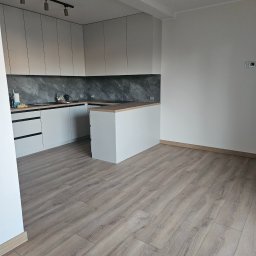 Soldatke Łazienki - Jasnoszara zabudowa kuchenna z ciemnym marmurowym blatem i drewnopodobną podłogą. Aneks kuchenny z wyspą, czarną szyną oświetleniową, w minimalistycznym stylu.