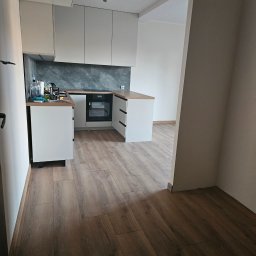 Soldatke Łazienki - Nowoczesna kuchnia z białymi szafkami, drewnianym blatem i ciemnym, marmurowym panelem ściennym. Wbudowany piekarnik, oświetlenie szynowe i jasne panele podłogowe w całym pomieszczeniu.