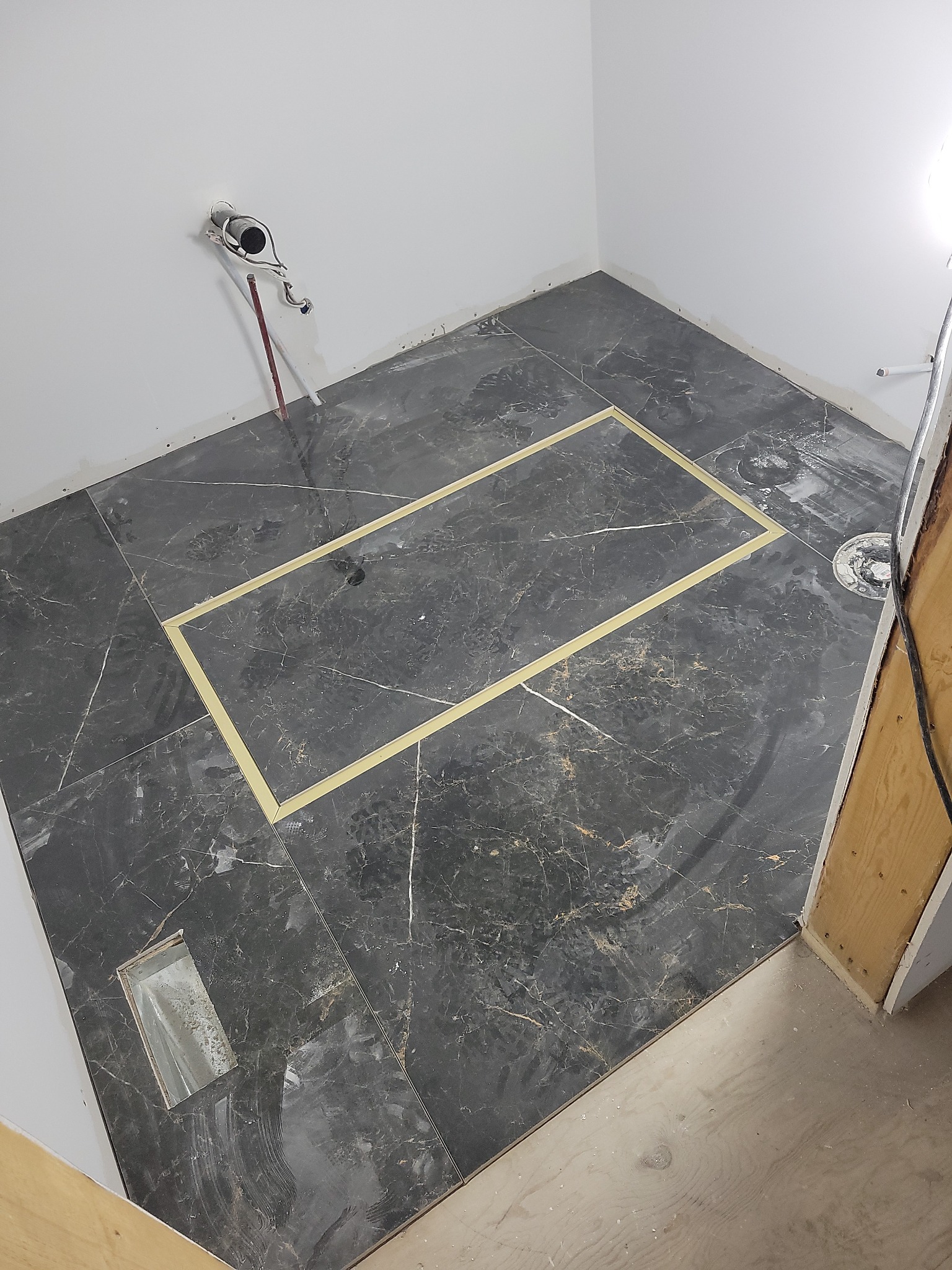 Ułożona podłoga z ciemnych płytek imitujących marmur w łazience. Widoczne taśmy wyznaczające obszar, przygotowane podejście wod-kan i odpływ liniowy.