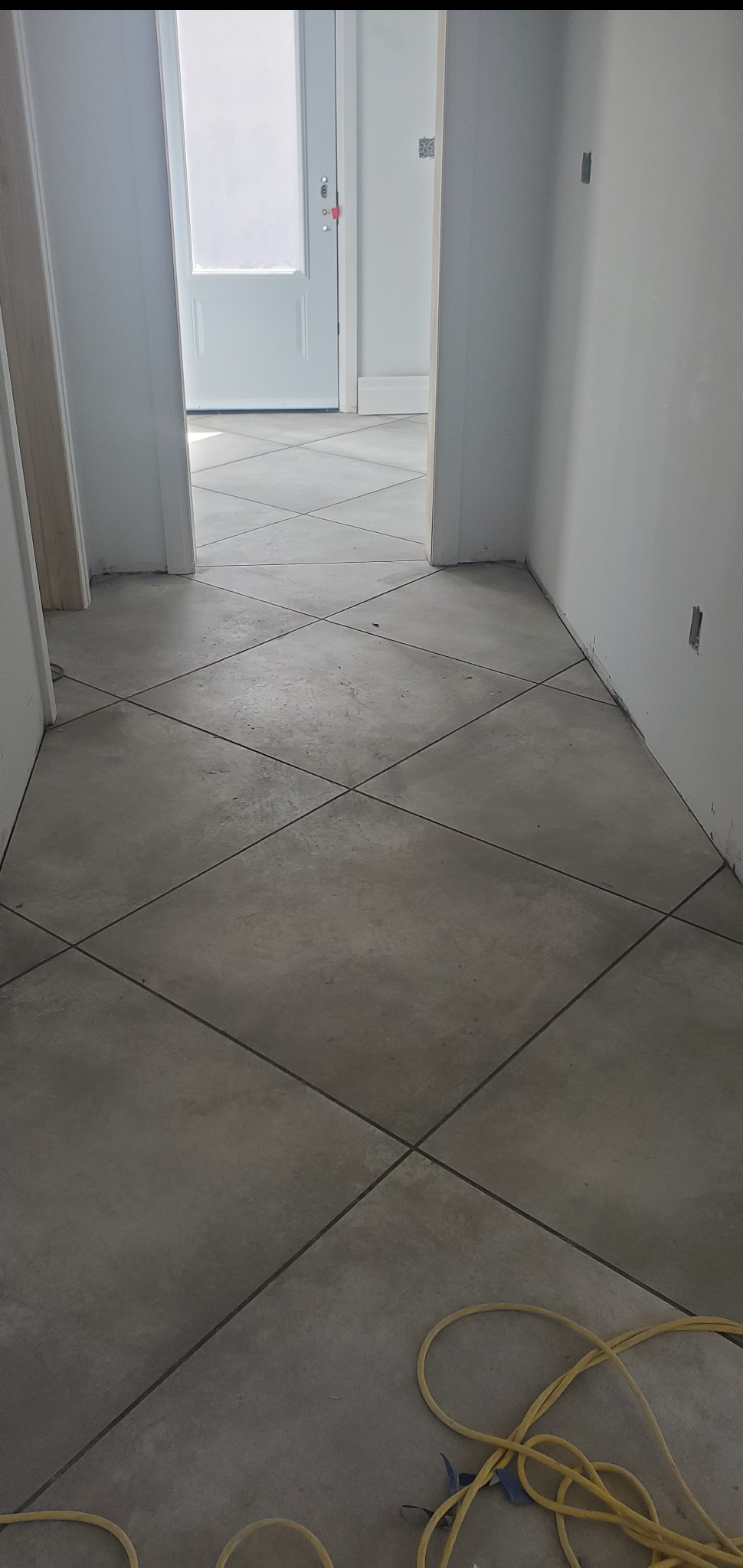 Ukończona podłoga z szarych, kwadratowych płytek ułożonych pod kątem. Widoczne przewody elektryczne. W tle białe drzwi i ściany w trakcie wykańczania.
