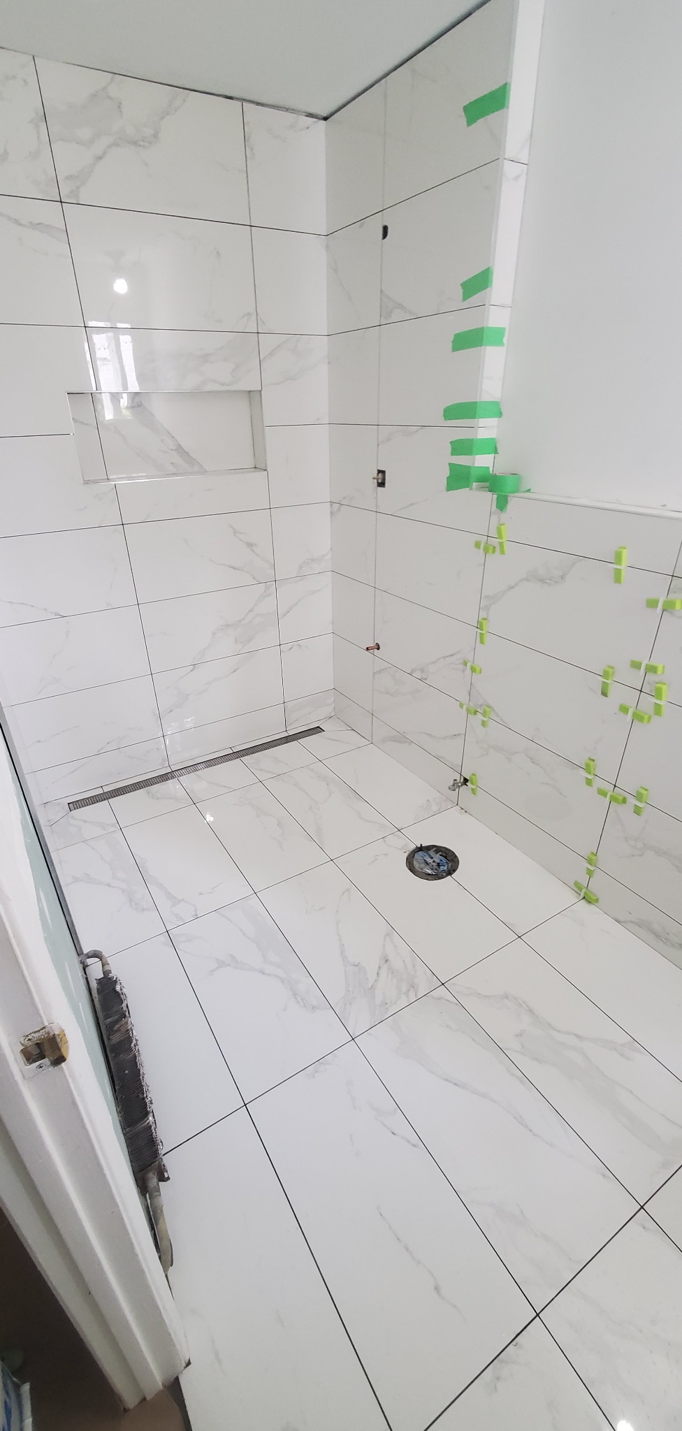 Łazienka w trakcie wykańczania: białe płytki imitujące marmur na ścianach i podłodze, zielone krzyżyki dystansowe i taśmy maskujące, odpływ liniowy.