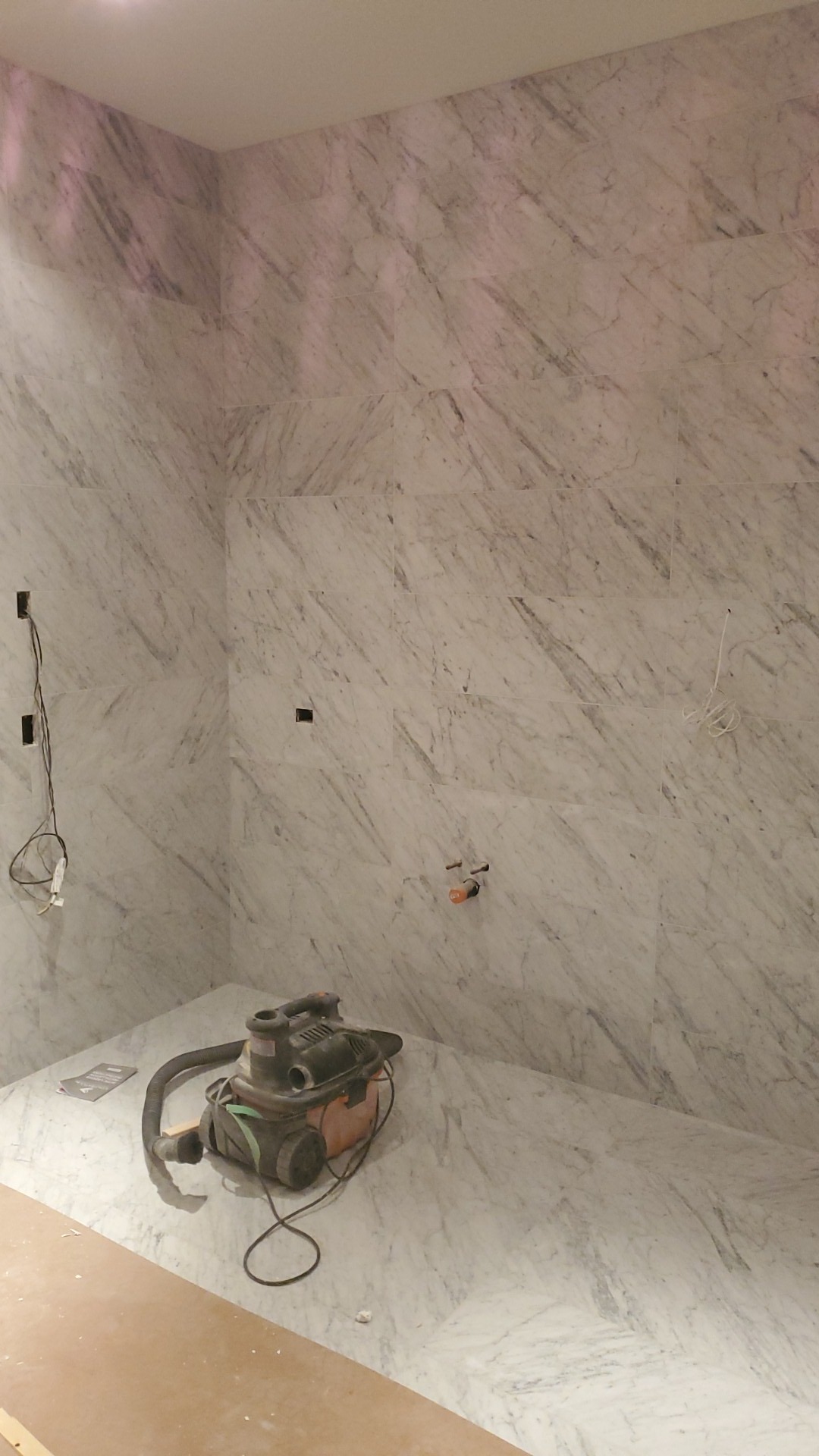 Łazienka w trakcie prac: marmurowe płytki na ścianach i podłodze, widoczne kable, rury i odkurzacz przemysłowy. Etap prac wykończeniowych.
