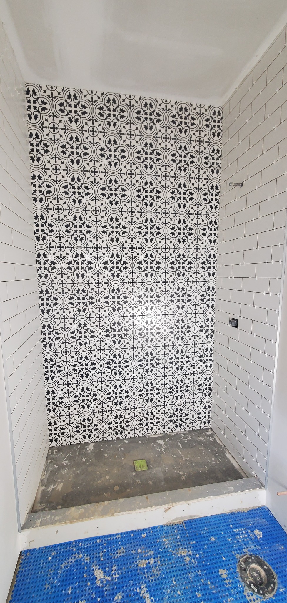 Ściany prysznica w trakcie wykańczania: białe, podłużne płytki na dwóch ścianach i dekoracyjne czarno-białe na trzeciej. Widoczny odpływ liniowy i niebieska mata izolacyjna.