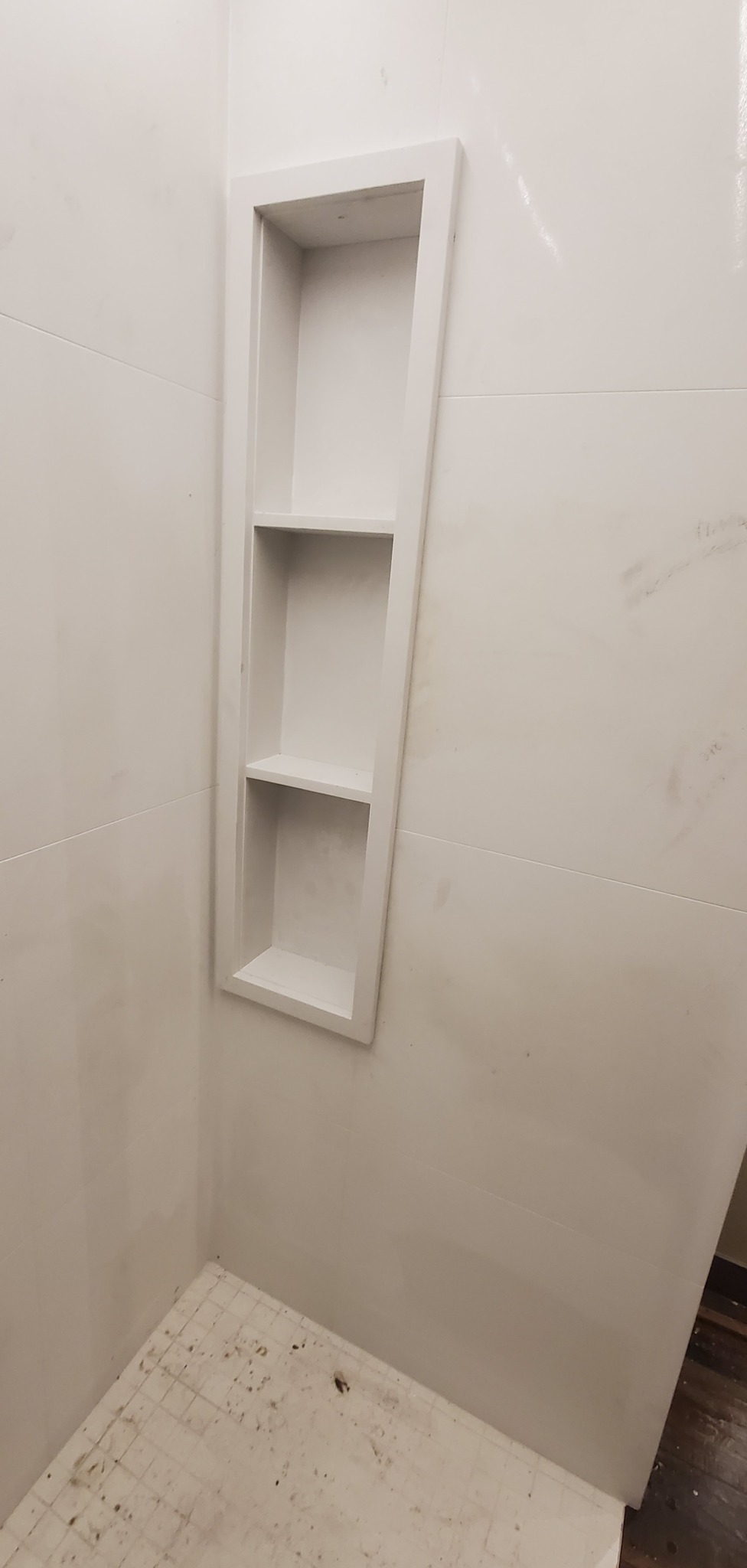 Biała, wnękowa półka z trzema poziomami w wykafelkowanej ścianie łazienki. Widoczny fragment podłogi z drobną mozaiką.