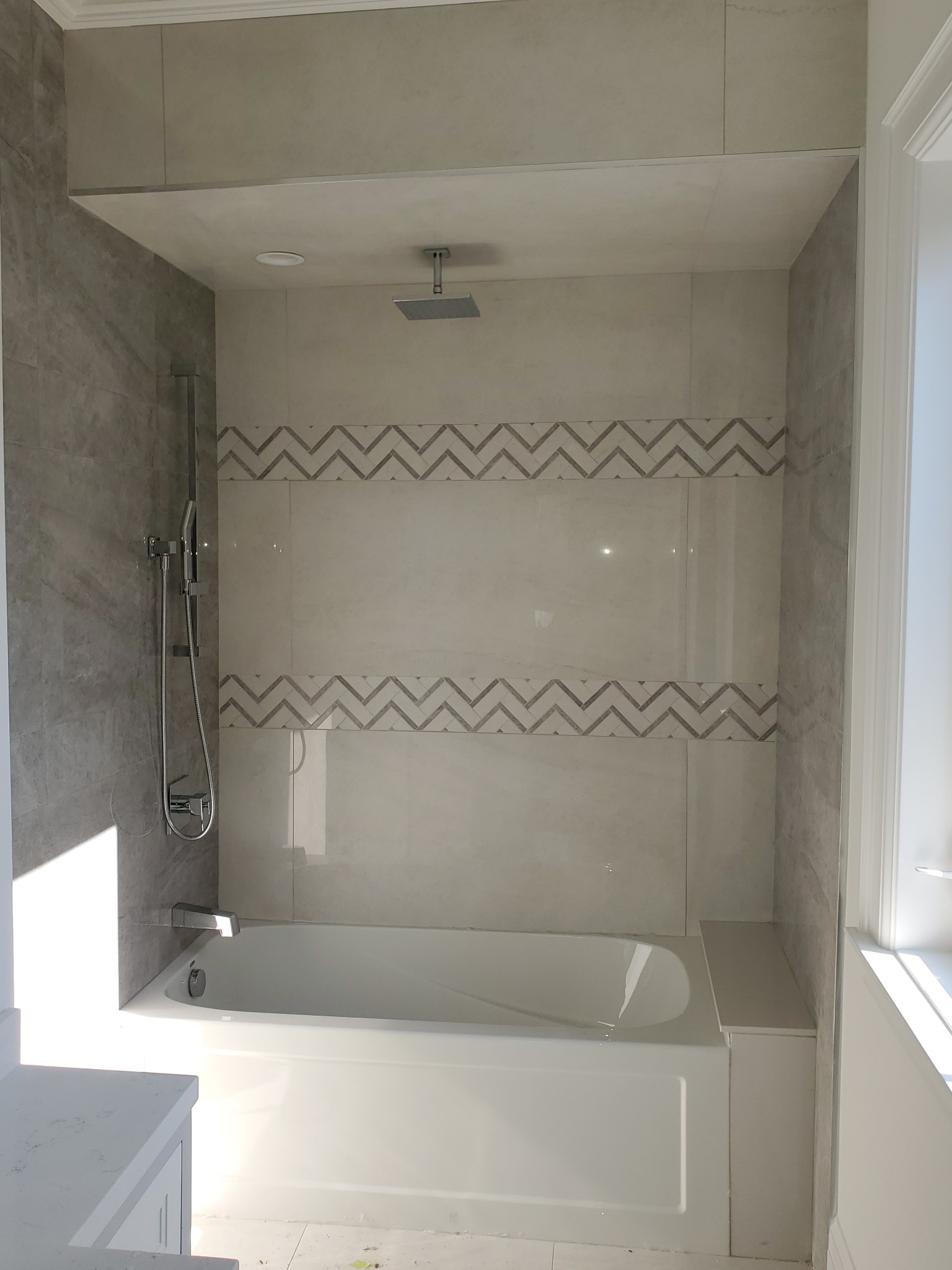 Wykończona łazienka: biała wanna pod prysznicem z szarą mozaiką w zygzaki na jasnych płytkach. Widoczny panel boczny wanny i chromowana armatura.