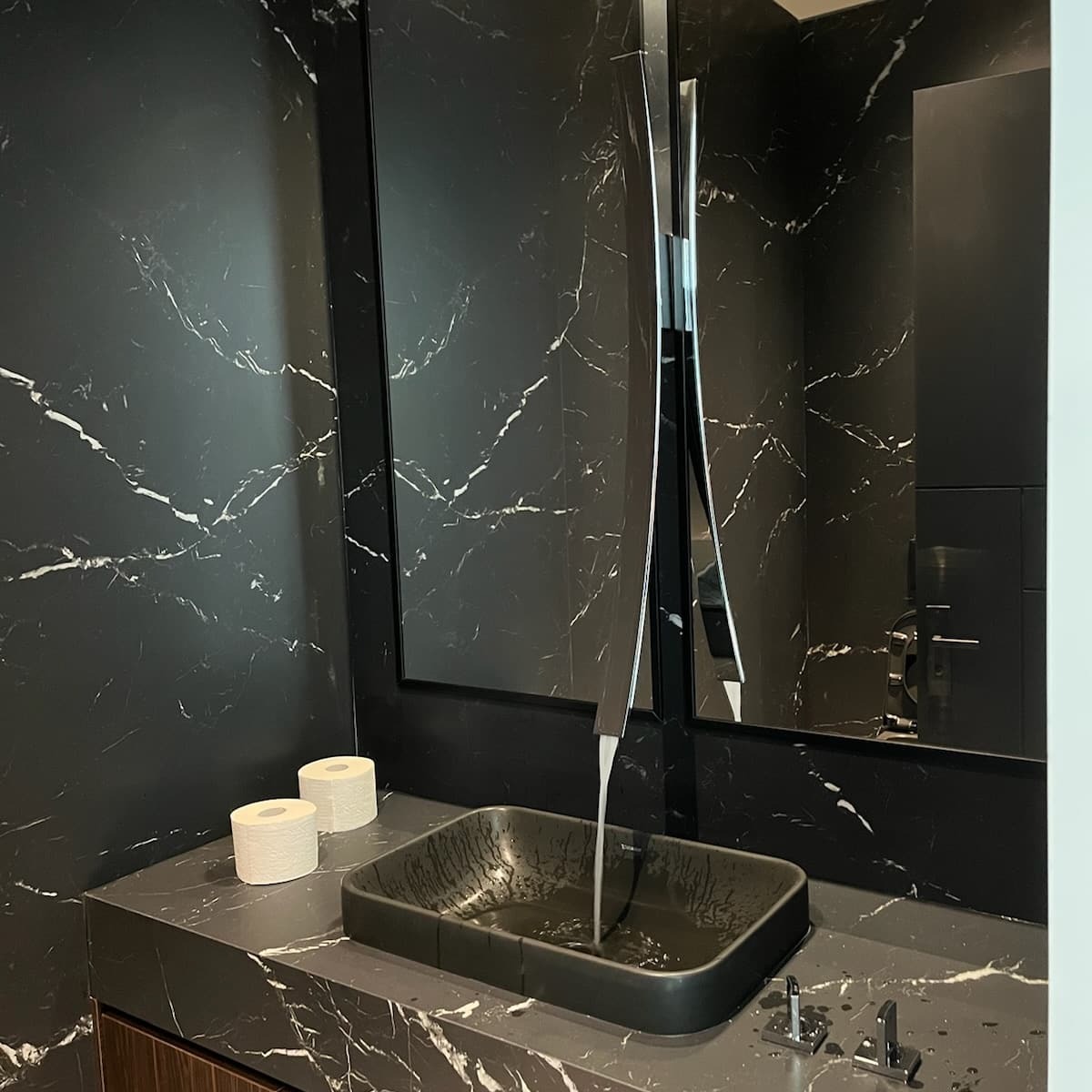 Elegancka łazienka z czarnym marmurem, nowoczesną umywalką i minimalistyczną armaturą. Widoczne dwie rolki papieru toaletowego i woda lejąca się z kranu.