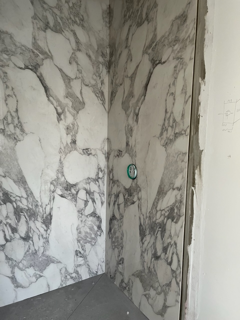 Wykończony narożnik łazienki z imitacją marmuru na ścianach. Widoczny fragment podłogi i instalacja. Na ścianie szkic z wymiarami.