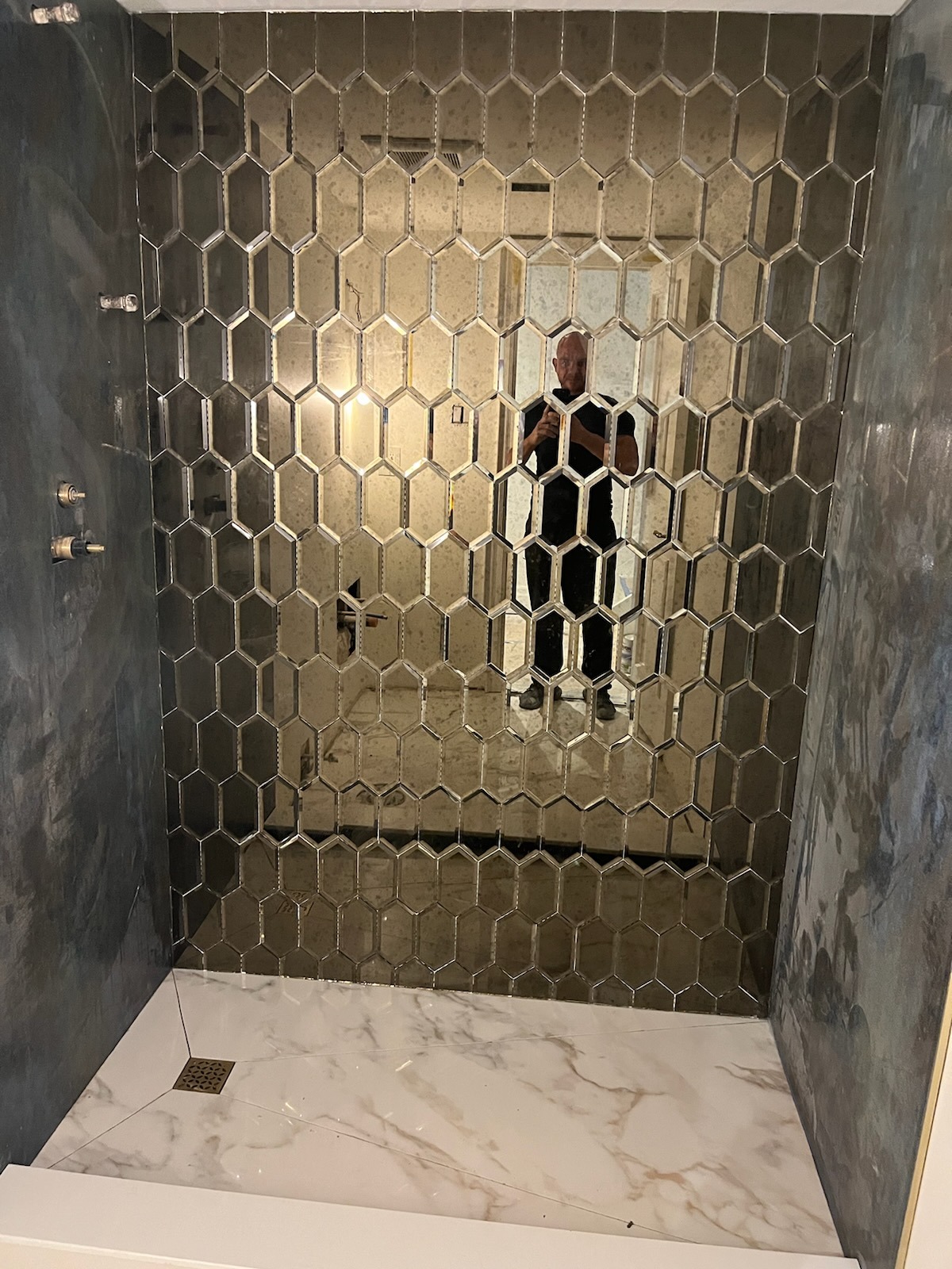 Wykończona kabina prysznicowa z heksagonalnymi, lustrzanymi płytkami na ścianie i marmurową podłogą z odpływem liniowym. Widoczne elementy armatury.